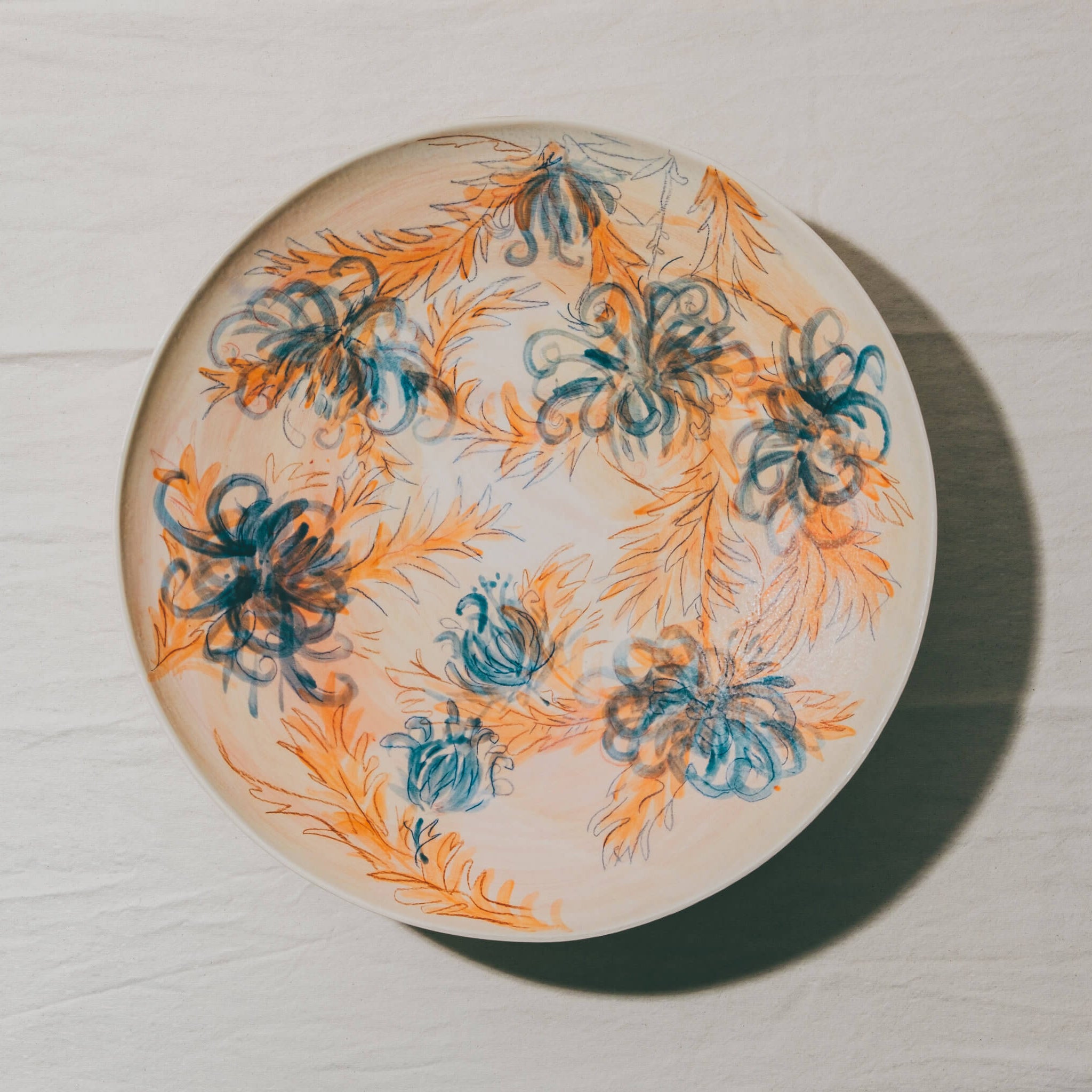 Plat coupe moulé à l’Atelier Grand Soleil, en faïence décorée au crayon à oxyde, à l’engobe puis émaillée transparent.
Les plats sont compatibles avec un lavage en machine en mode éco.
Possibilité de personnalisation à l'or et à la plume. 
Motif : chrysanthèmes, dans les tons orange et bleu